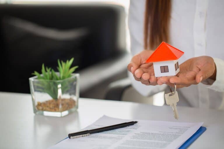 A house figurine and key on the hands of a woman; a contract in the background in the concept of moving houses