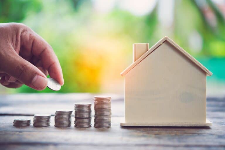 A cropped photo of a man's hand stacking coins with a wooden house figurine in the concept of saving money when moving to Ilford.